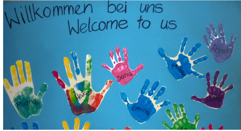 Auf einem blauen Hintergrund sind bunte Hände mit Namen darin abgebildet. Oben drüber steht auf Deutsch und Englisch: Willkommen bei uns.